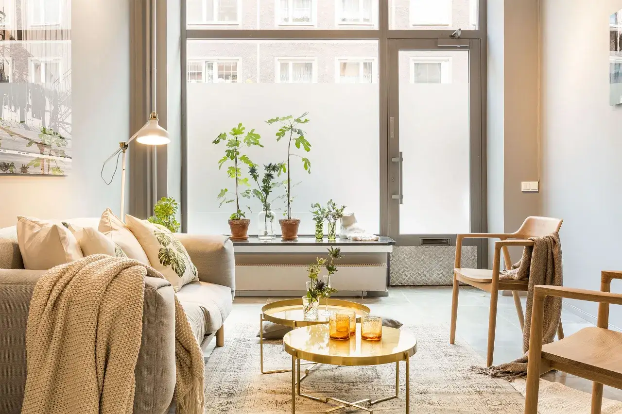 Un salon baign&eacute; de lumi&egrave;re naturelle, avec un canap&eacute; confortable, des tables basses dor&eacute;es et des plantes vertes, cr&eacute;ant une atmosph&egrave;re de feng shui.