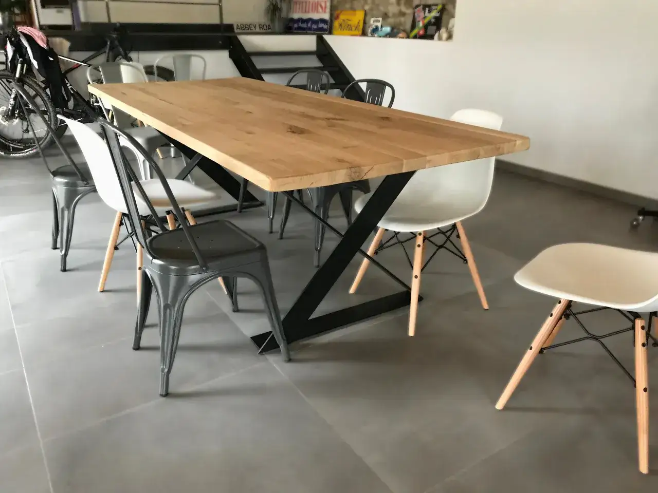 Un grand plateau en bois repose sur un piètement noir, entouré de quelles chaises avec table industrielle. Des chaises en métal gris et des chaises blanches modernes complètent l'ensemble.