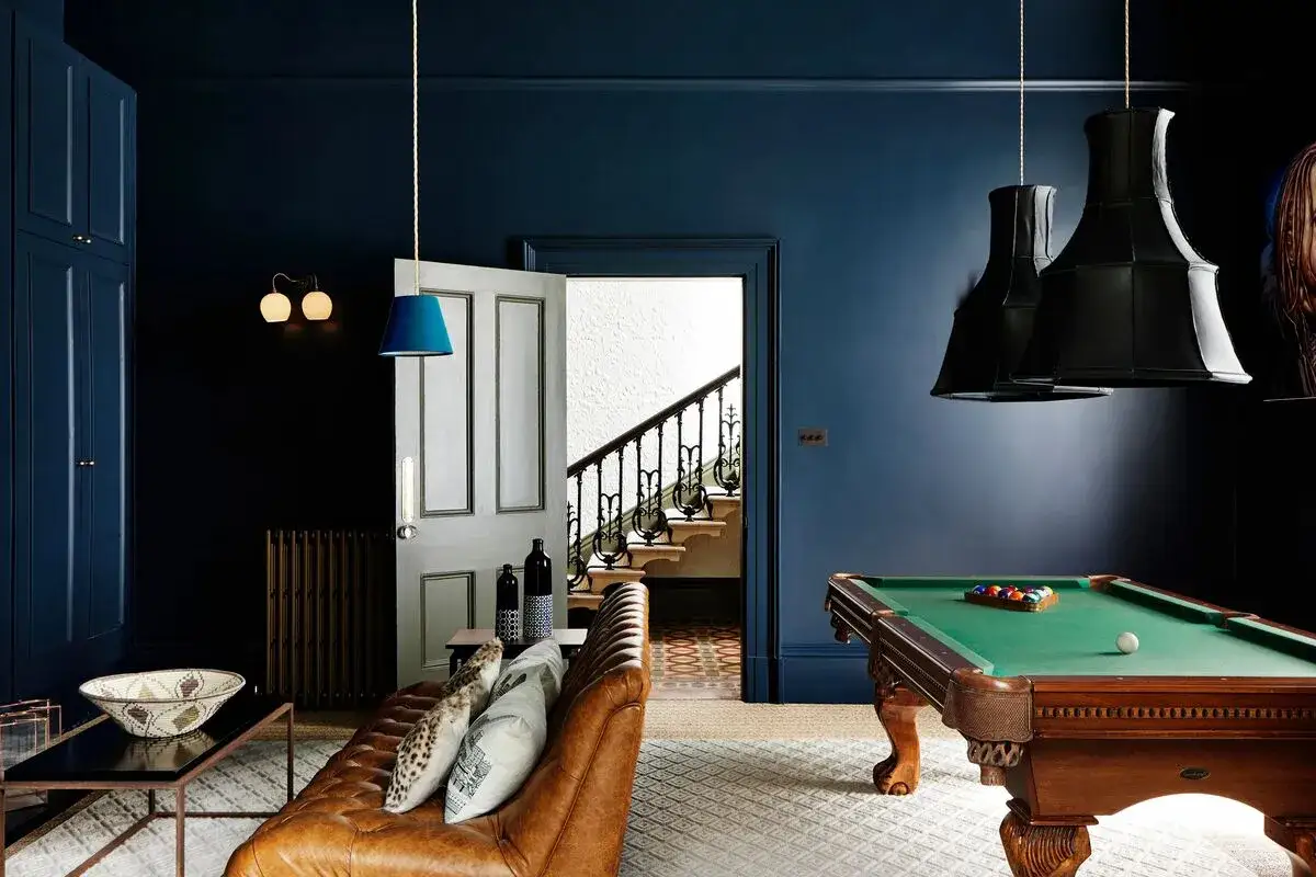 Salon avec billard et canap&eacute; en cuir marron, murs d'un profond couleur bleu fonc&eacute;. Une porte ouverte r&eacute;v&egrave;le un escalier.