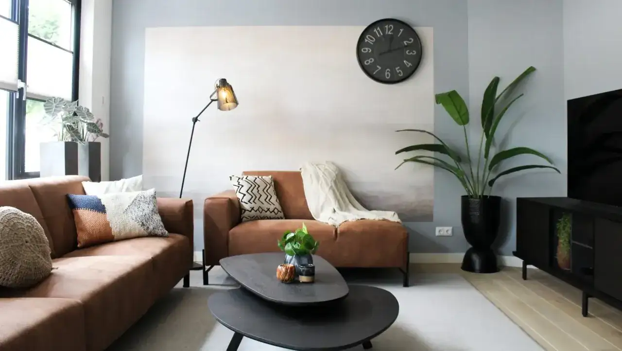 Quel budget pour meubler une maison neuve ? Salon moderne avec canap&eacute; marron, coussins d&eacute;coratifs, table basse, plante verte et horloge murale.
