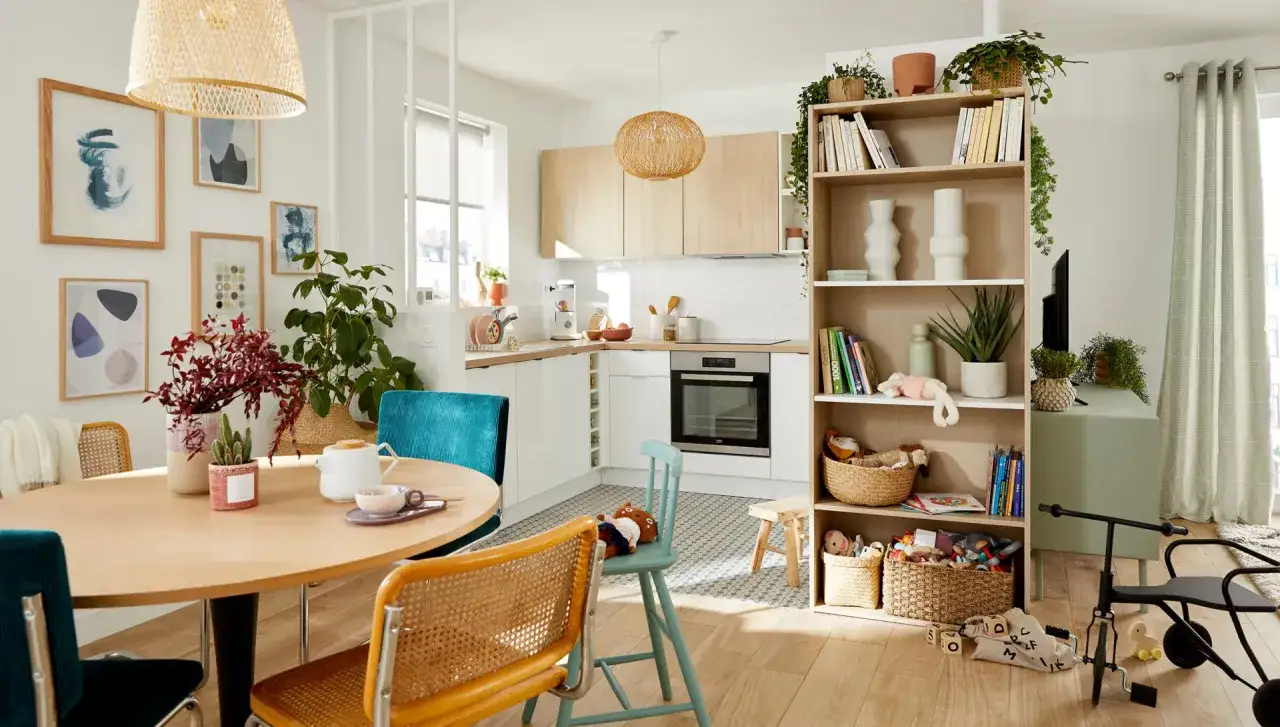 Une petite salle &agrave; manger lumineuse avec une table ronde en bois, des chaises color&eacute;es, une cuisine moderne et une &eacute;tag&egrave;re remplie de livres et de jouets.