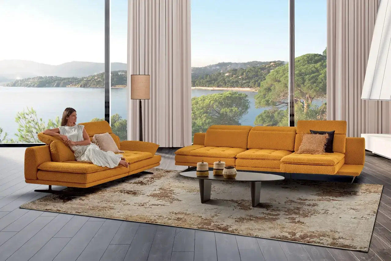 Une femme se détend sur un canapé jaune moutarde, profitant d'une vue imprenable sur la mer. Ce canapé, conçu pour une longue durée de vie, s'intègre parfaitement à ce salon lumineux.