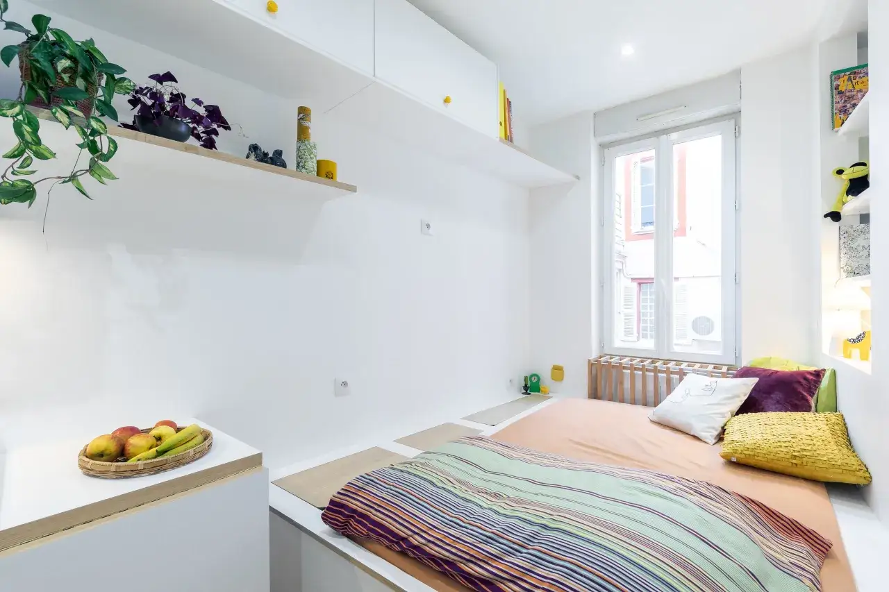 Chambre avec lit, coussins colorés, plantes et fruits. La position bz du lit est optimisée pour l'espace.