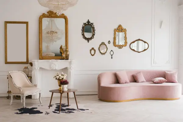 Un salon élégant avec un canapé rose, une chaise vintage, une cheminée ornée et une collection de miroirs dorés. Une déco salon vintage moderne réussie.